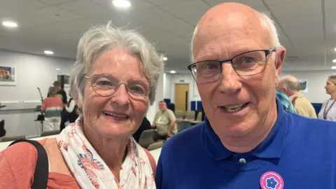 George Carden/BBC Peter wearing glasses and a blue jumper and polo top, his wife Wendy has gray hair and wearing a peach coloured top and has a white scarf on