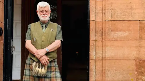 Anna Holligan James MacLeod, concierge at the Glencoe House Hotel, stands outside the entrance, wearing traditional Scottish attire, including a kilt