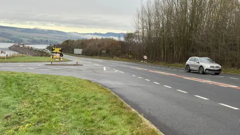 BBC A junction on the A9 with the Cromarty road bridge and Cromarty Firth in the distance. A silver SUV is passing the junction.