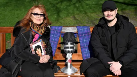 Greg Macvean A woman with copper hair wearing sunglasses and wearing a black coat and patterned scarf she is holding an old photo of her husband, Les McKeown. She is sitting on a bench next to a man who wearing all black. Between them is a microphone with a hat placed on it