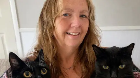 A middle-aged woman with blonde hair smiles at the camera. Two black cats are visible in front of her.