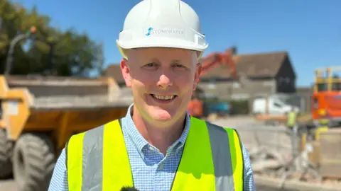 A man in a white hard hat and yellow high-vis vest over a blue checked shirt standing on a building site smiling at the camera on a sunny day