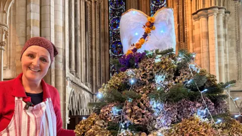 Karen Simpkin - a woman in a red top, with a red and white striped apron on and a scarf on her head. She is standing next to a Christmas tree in the minster with dozens of petals and flowers attached to it. 