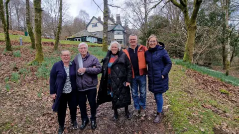 Alan Kelly stands with his family members, two women and a man, in front of Hawes End near Keswick. The Victorian building is made up of various sections, with numerous chimney pots on the pitched roof. It sits on top of a slope, which is covered in leaves and surrounded by birch trees.