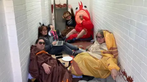 A group of five people wearing inflatable animal costumes - including a lobster and a monkey costume - sit on the floor of a brick kennel at animal shelter.