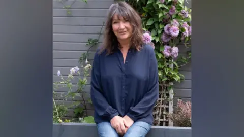 Handout A woman in a blue shirt and jeans sits on a garden wall with flowers behind her