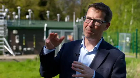 Prof William Bloss wears a navy suit and open collar pastel blue shirt and dark rimmed glasses. He is pictured gesticulating with his hands as he talks outside the university's monitoring station on campus which takes readings of nitrogen dioxide and particulate matter 