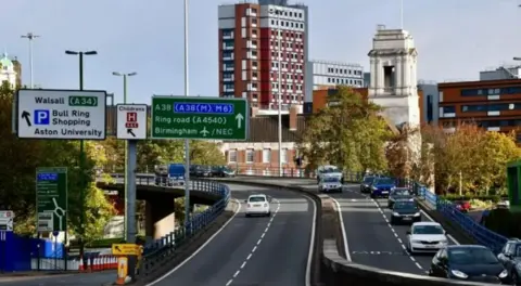 A flyover section of the A38 Aston Expressway on the way into Birmingham city centre. On the left there are a series of road signs, indicating the likes of Walsall and parking for Bull Ring Shopping. Vehicles can be seen on both carriageways, with buildings in the distance. It is a sunny day.