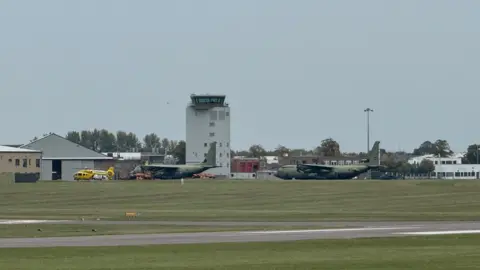 Steve Hubbard/BBC Several aircrafts are on an airport which has a runway in the foreground. In the distance in the middle is an air traffic control tower.