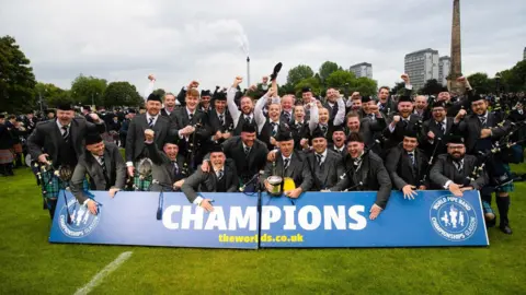 World Pipe Band Championships Members of a pipe band celebrate being crowned world champions. About 20 members dressed in kilts and jackets look happy posing behind a blue champions banner.