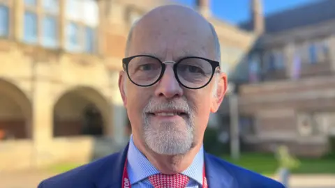 A man with glasses and a white beard smiles at the camera. He is standing in front of the school and wears a blue suit jacket, blue stripe shirt and red checked tie. He is wearing a red lanyard.