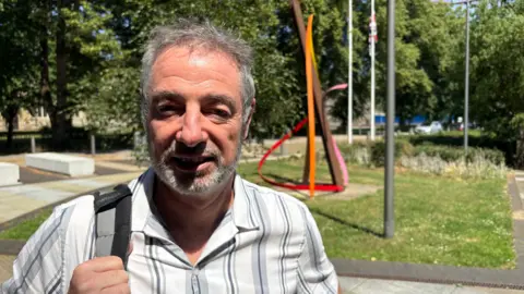 Martin Heath/BBC David Hewson, with short white hair and beard, standing outdoors in a park, wearing a striped shirt and holding a shoulder strap, with trees, flagpoles and a colourful abstract sculpture in the background on a sunny day.