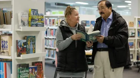 A woman and man stand next to library shelves and hold a book between them, discussing it. The woman has a pony tail and is wearing a black gillet, grey hoodie and black leggings. The man has black hair and is wearing a winter coat, blue shirt and cream chinos.