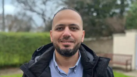 A man with dark hair and a short beard smiles at the camera, He is outdoors, possibly in a garden, and wears a light blue shirt and a dark rain jacket.