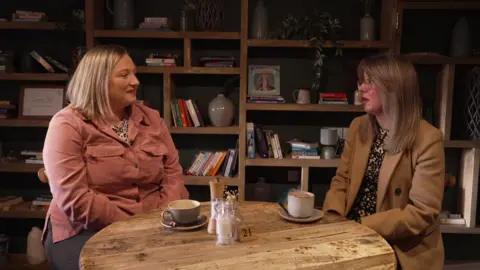 The photo shows two women seated at a small round wooden table inside what appears to be a cosy café or coffee shop. The table has a rustic, worn texture and holds two cups of coffee, a small jar of sugar, a salt and pepper set, and a wooden table marker with the number 21.
Behind them is a large wooden shelving unit that fills the background. The shelves contain a mix of books, framed pictures, small decorative vases, plants, and ornaments arranged in a casual, homely style. 