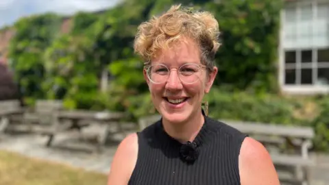 Philippa Knott Kos is wearing glasses and a black ribbed sleeveless top and she is standing in front of picnic benches and a wisteria-covered building.