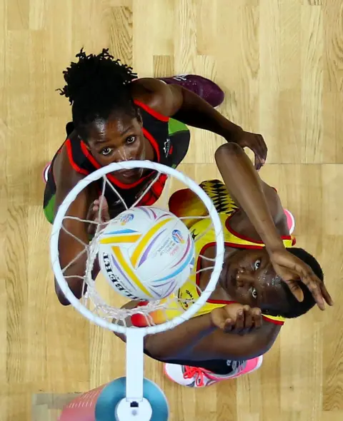 PA Two Netball players stand next to the net, watching the ball go in. They are both reaching upwards and looking intently at the ball.