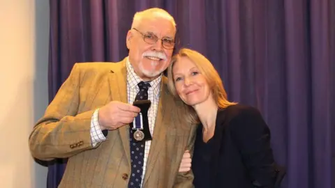 Joe Ousalice A full-colour picture of a man showing a military medal as a woman leans on his left shoulder