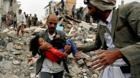 Reuters A man carries a girl from the site of a Saudi-led coalition air strike in Sanaa, Yemen (25 August 2017)