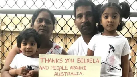 HOMETOBILO/TWITTER A family picture of Nadesalingam and Priya and their two daughters holding a sign reading "thanks you Biloela and people around Australia"