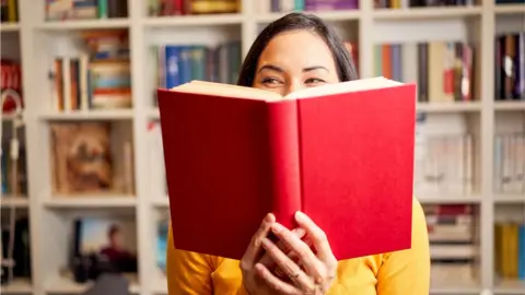 Getty Female young behind book with face covered for a red book while smiling - stock photo