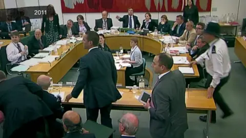 UK Parliament Emily Commander was clerk of the Culture, Media and Sport Committee when Rupert Murdoch was attacked with a foam pie. She is on the right side of the chair, John Whittingdale, the man at the centre of the table with his arm raised. Paul Farrelly is seated three seats away further to the right.