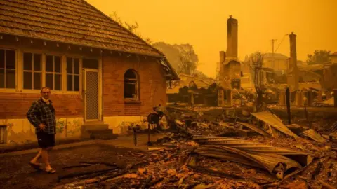 Getty Images A man stands in front of burnt-out buildings in Cobargo, New South Wales, below an orange sky filled with smoke