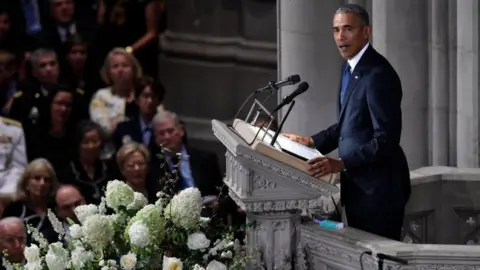 AFP/Getty Images Former US President Barack Obama speaks at the memorial service, from a lectern flanked by white flowers