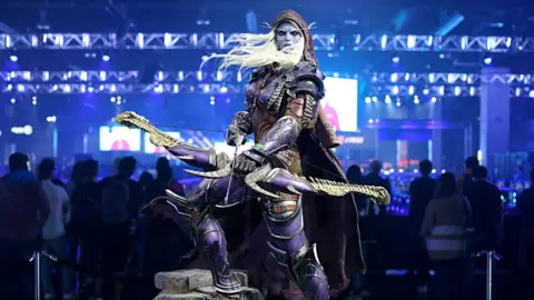 Getty Images A statue of a character from World of Warcraft with purple skin and pointy ears holding a bow and standing in front of a crowd in a big room with bright lights