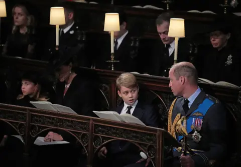 PA Media (front row, left to right) Princess Charlotte, the Princess of Wales, Prince George, and the Prince of Wales, watch as the Imperial State Crown and the Sovereign's orb and sceptre are removed from the coffin of Queen Elizabeth II, draped in the Royal Standard, during the Committal Service at St George's Chapel in Windsor Castle, Berkshire