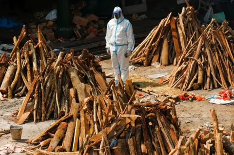 Adnan Abidi / Reuters A man wearing personal protective equipment (PPE) stands next to funeral pyres in New Delhi, India