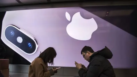 Getty Images Two people look at their iPhones in an Apple store, dwarfed by an enormous promotional image for the latest model