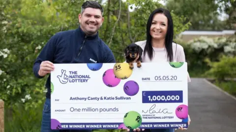 PA Media Anthony Canty, Katie Sullivan and Lucky Lola the dachshund with a giant cheque