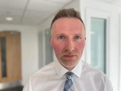 Jason has short cropped hair and a beard. He is wearing a shirt and tie and is stood in an office.