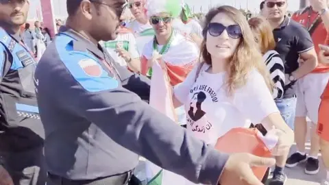 A woman wearing a Mahsa Amini t-shirt being obstructed by a police officer