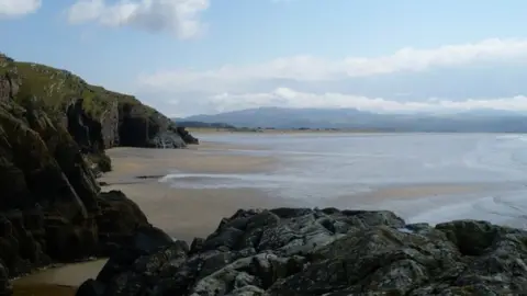 Geograph/Chris Carlson  black rocks above a long sandy beach with waves and mountains in the distacee