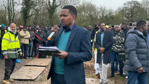 A man speaking into a microphone with a large crowd in the background