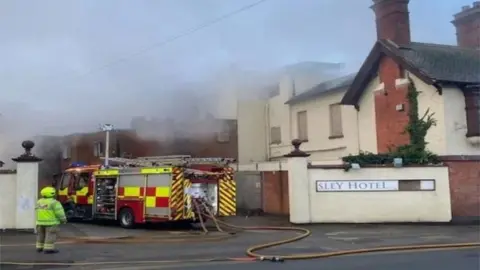 West Midlands Fire Service Fire engine outside a building with boarded up windows and plumes of smoke visible