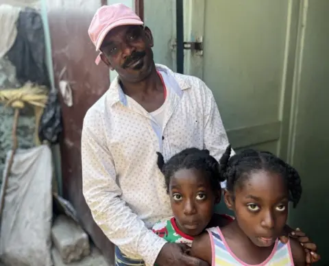 A moustached man in a pink hat smiles at the camera, his hands on the shoulders of his young daughters