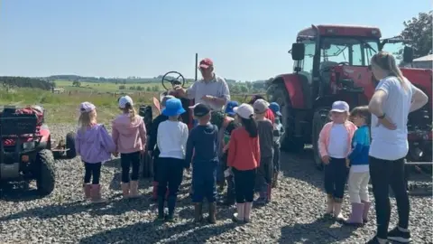 The Country Trust  A group of children gather round Charlie Bennett with a teacher watching and a tractor in the background