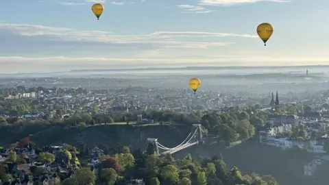 Three hot air balloons over Bristol with the Clifton Suspension Bridge in the foreground on a sunny day