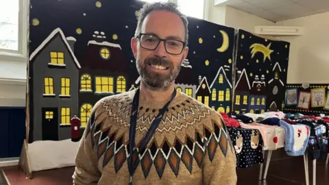 Stephen Morton. He has glasses and facial hair. He is wearing a beige Christmas jumper with patterns across the shoulders. He is smiling at the camera. Behind him, a Christmas display can be seen, as well as a collection of jumpers laid out on a table.