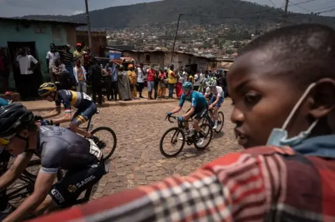 AFP A child looks on as he watches cyclists competing during the final stage of the 14th Tour du Rwanda on 27 february 2022, in Kigali. - Eritrean Natnael Tesfatsion won the Tour of Rwanda for the second time, which ended on Sunday in the capital Kigali with the victory of Rwandan Moïse Mugisha in the 7th stage.