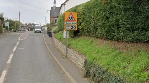 Google A yellow sign by the side of the street with a red triangular school crossing warning, and the wording "School Zone - Max Speed 20". 