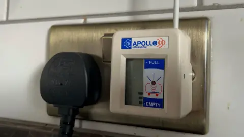 Ellen Knight/BBC Close-up of a fuel gauge, which is a white box plugged in to a gold, shiny wall unit. The bars on the fuel gauge are low, with only three visible, about a quarter of the way up. There is a blue label with white writing at the top that reads 'Apollo ultrasonic', and to the right of the bars indicating how full it is there is another label with 'full' at the top and 'empty' at the bottom. 