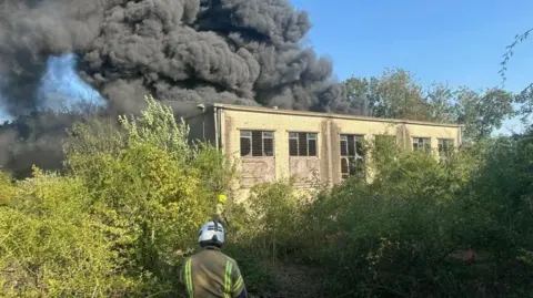 ECFRS Fire officer in protective clothing and helmet stands in front of a pale -coloured one storey building. Smoke billows from the back of the unit. Shrubbery surrounds the site.