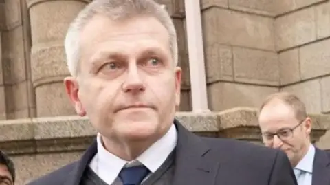 Deputy Philip Ozouf is outside the Royal Court. He is wearing a black suit with a blue tie.