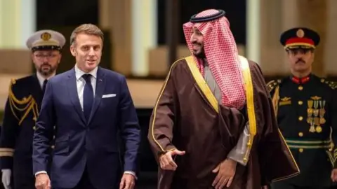 Shutterstock Emmanuel Macron walks alongside Saudi Crown Prince Mohammed bin Salman (R) at Al-Yamamah Palace in Riyadh, Saudi Arabia, with two men in military uniform walking behind them.