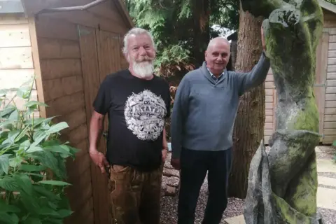 Geraldine O'Malley Simon Gee in a black t-shirt with a white image on it stands in a garden next to George Wagstaffe. George has on a blue jumper and dark blue trousers and has his hand on one of his sculptures. 