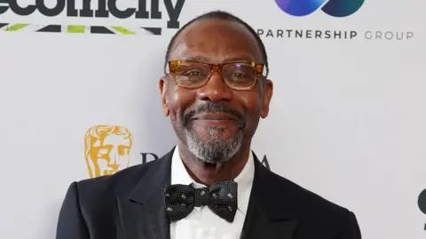 Lenny Henry smiles at the camera as he attends an awards ceremony. He is wearing brown glasses as well as a black suit, bow tie and white shirt. 